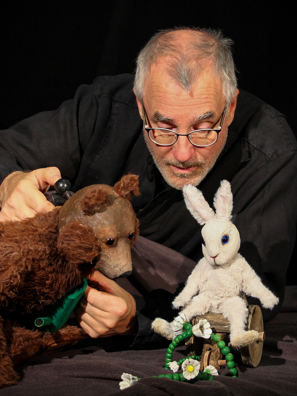 Ein Puppenspieler mit Brille hält eine braune Bärenfigur, die zu einem weißen Hasen auf einem kleinen Wagen blickt. Der Hintergrund ist schwarz, und vor dem Hasen liegen dekorative Blumen.