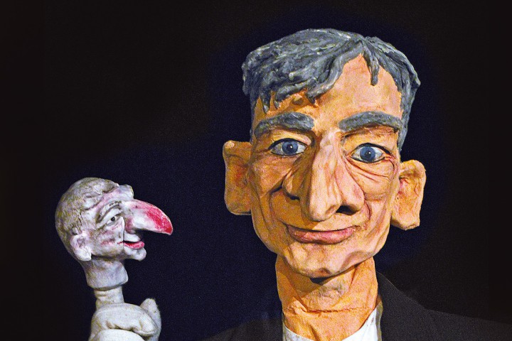 Eine Marionette mit markanten Gesichtsz&uuml;gen und kurzen grauen Haaren tr&auml;gt einen schwarzen Anzug. Daneben eine kleinere, wei&szlig;e Figur mit spitzer Nase, die in ihre Richtung blickt, vor schwarzem Hintergrund.