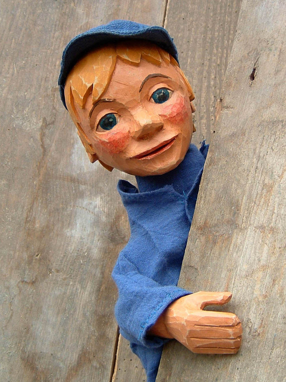 Holzfigur eines Jungen mit blonden Haaren, blauen Augen und rotem Gesichtsausdruck, in blauer Kleidung und Mütze, schaut lächelnd hinter einem Holzbalken hervor.