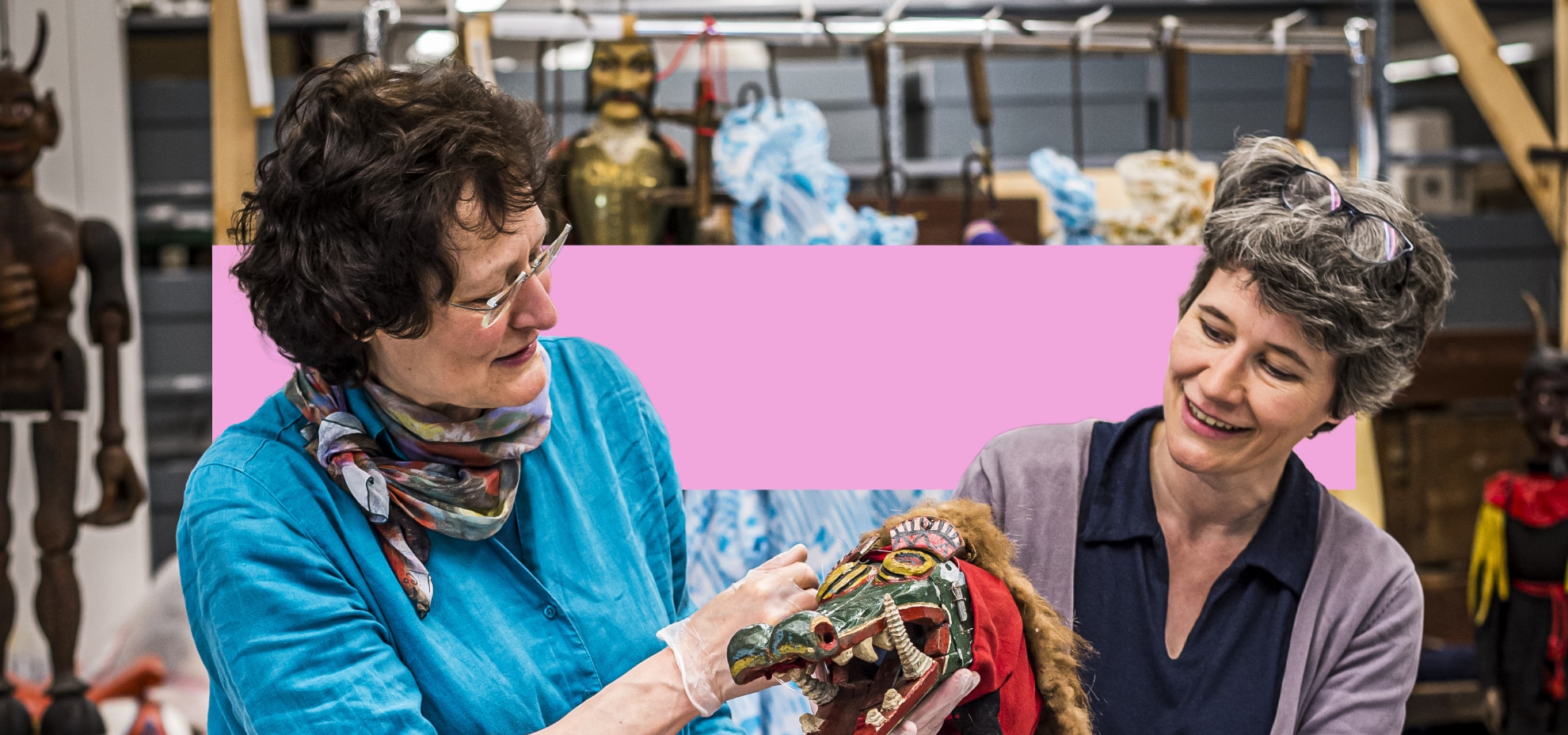 Zwei Frauen in einem Lagerraum für Theaterfiguren betrachten eine kunstvoll geschnitzte, farbenfrohe Maske mit tierischen Zügen. Im Hintergrund hängen weitere Figuren und Requisiten.