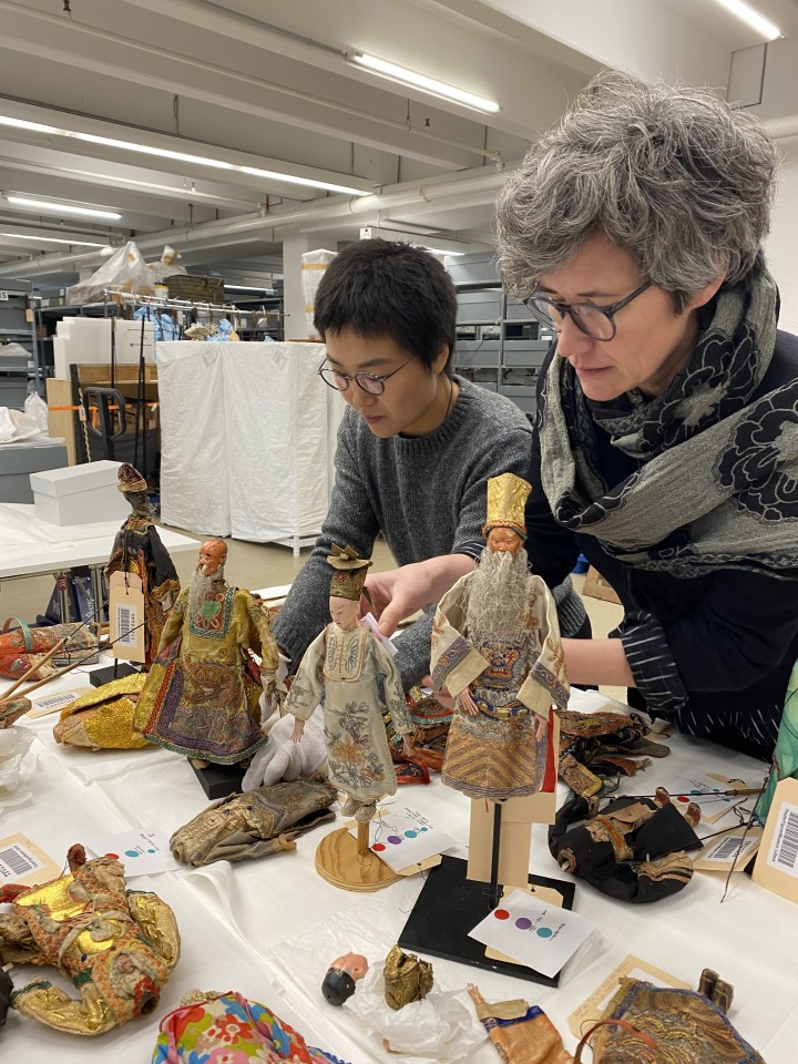 Zwei Personen untersuchen kunstvolle chinesische Figuren mit detaillierten Gewändern in einem Lagerraum. Der Tisch ist mit weiteren Puppen, Stoffen und Archivmaterial bedeckt