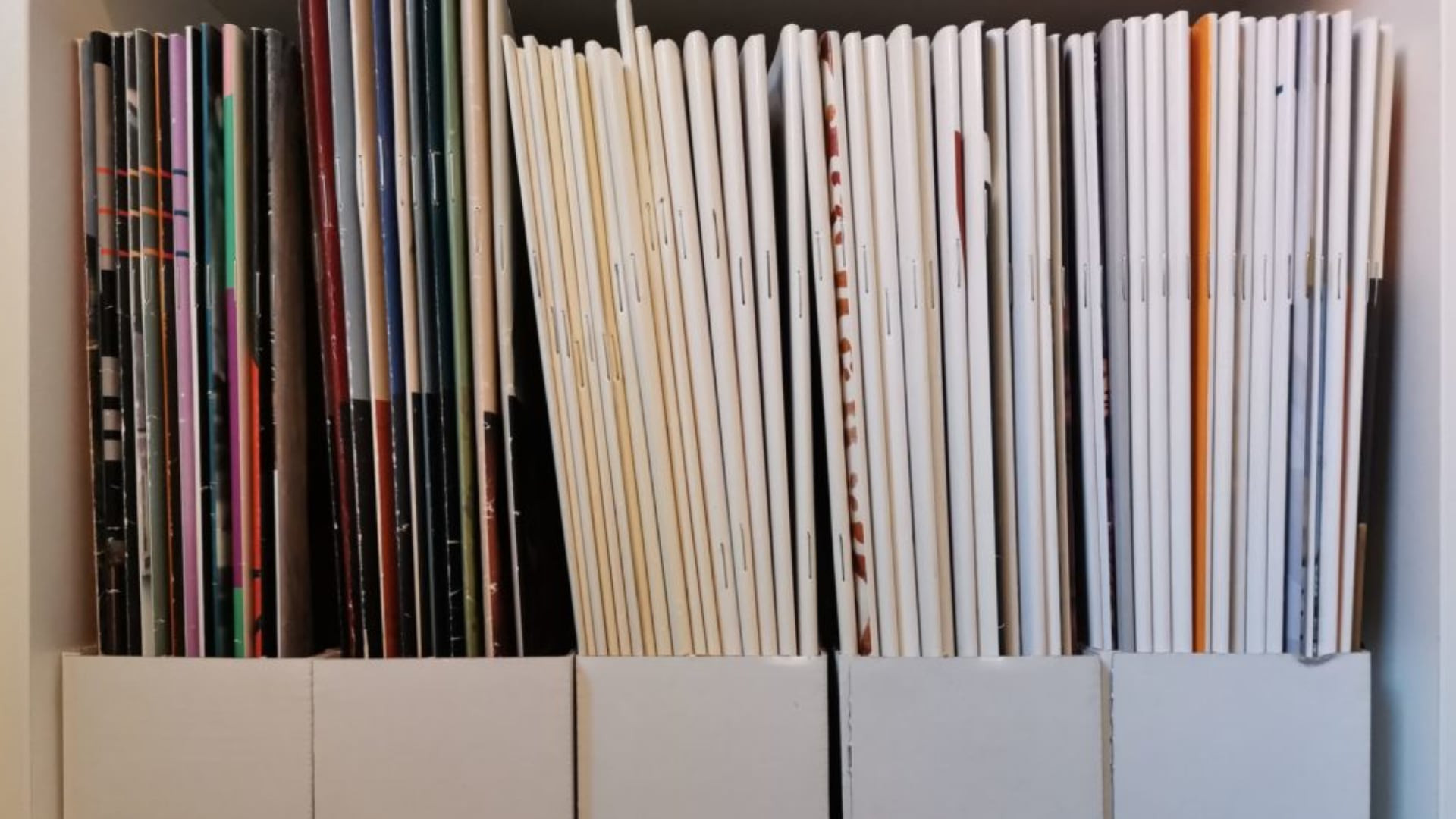 Nahaufnahme einer geordneten Sammlung von Heften und Magazinen in weißen Archivboxen, aufrecht stehend in einem Regal.