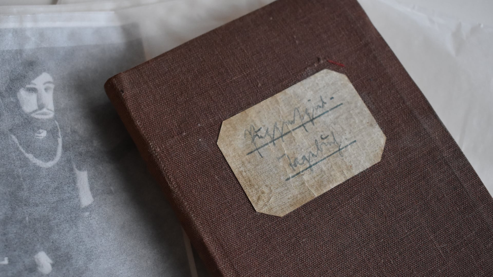 Nahaufnahme eines alten, braunen Stoffeinbandbuchs mit handgeschriebenem Etikett. Daneben liegt ein vergilbtes Foto oder Dokument mit dem Bild einer Figur unter Transparentpapier