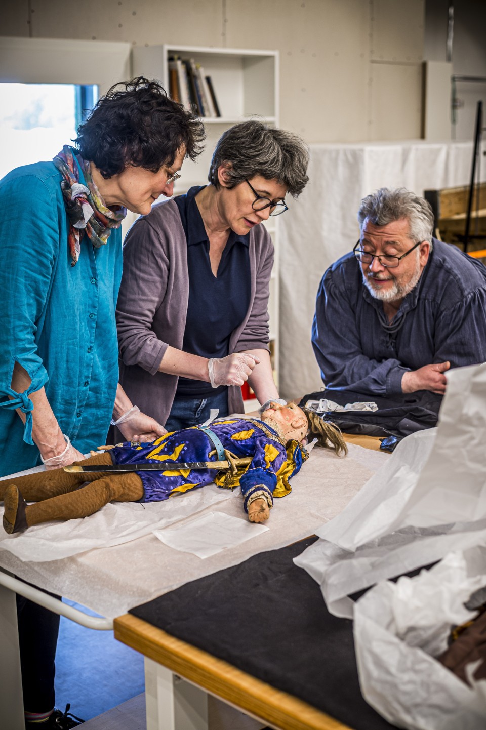 Drei Personen untersuchen eine historische Puppe in aufwendiger blau-goldener Kleidung auf einem Tisch. Sie tragen Handschuhe und arbeiten sorgfältig in einem Archiv- oder Restaurierungsraum.
