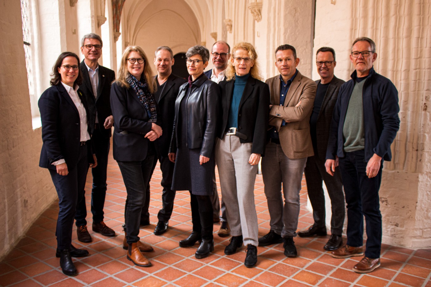 Eine Gruppe von zehn Personen in Business- und Smart-Casual-Kleidung steht in einem historischen Gebäude mit gotischen Bögen und Backsteinboden. Sie blicken freundlich in die Kamera.