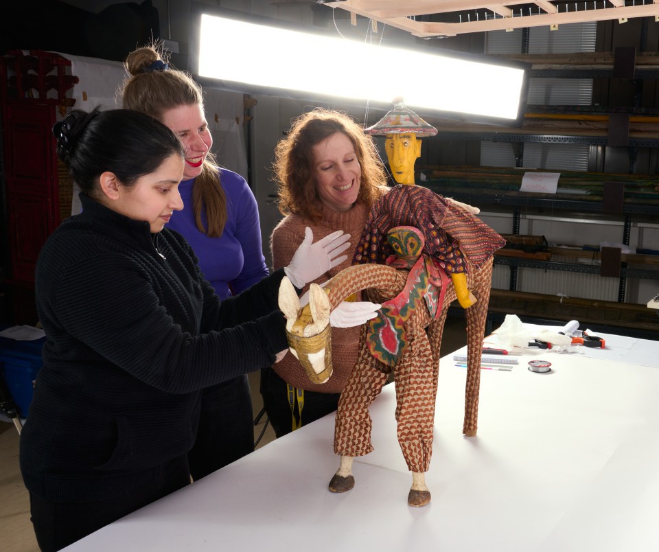 Drei Frauen untersuchen und restaurieren eine kunstvoll gestaltete, farbenfrohe Figur mit tierischen und menschlichen Elementen. Sie tragen Handschuhe und arbeiten an einem weißen Tisch in einem gut beleuchteten Archivraum.