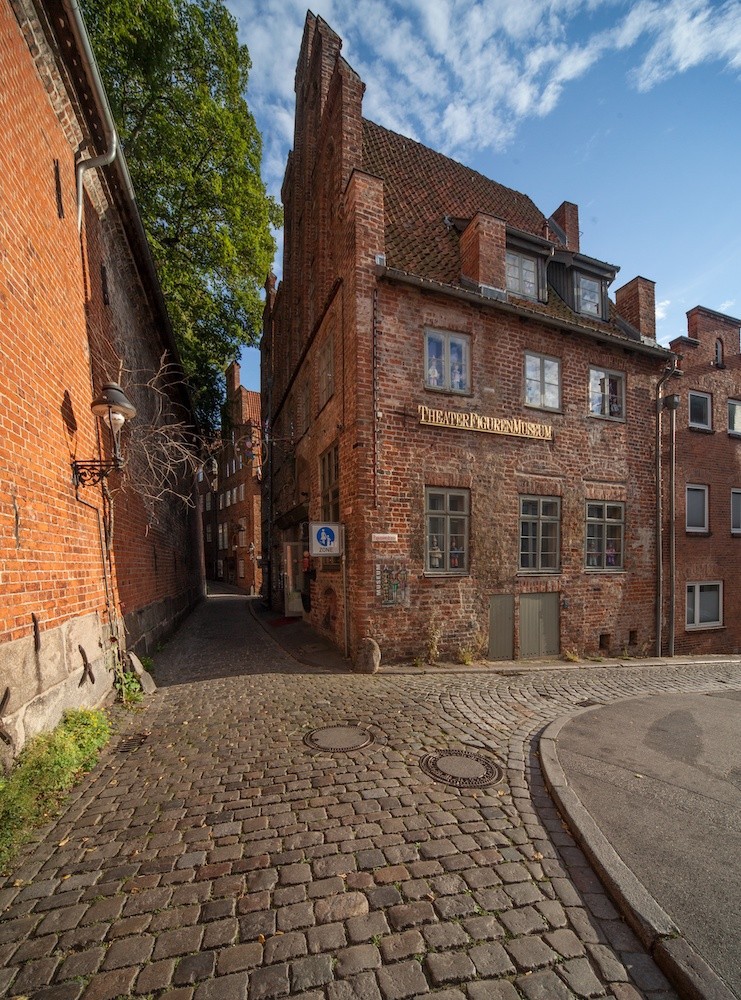 Das Bild zeigt das TheaterFigurenMuseum, ein historisches Backsteingebäude mit norddeutscher Backsteingotik. Es steht an einer Kopfsteinpflasterstraße und wirkt mit seinem warmen Lichteinfall malerisch.