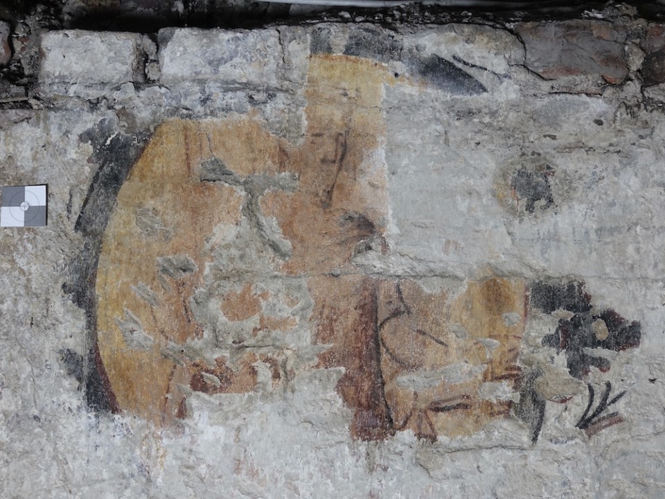 Die Wandmalerei zeigt verblasste, historische Motive mit erkennbaren Gesichts- und Pflanzenfragmenten. Der Erhaltungszustand ist stark beeinträchtigt, doch Farbspuren und Konturen lassen eine kunsthistorische Bedeutung vermuten.
